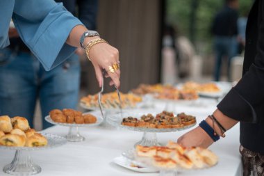 Guests at an elegant outdoor reception are choosing delicious appetizers and savory finger foods from a buffet table decorated with white tablecloths and crystal serving dishes