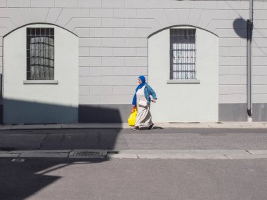 Cremona, Italy - September 18th A woman wearing a blue hijab and a patterned dress walks down a sunny street in Italy carrying a bright yellow bag past a building with arched windows and barred openings.