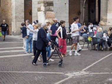 Cremona, Italy - September 18th 2025 Elderly couple with walking frame navigates a cobblestone square near historic Italian architecture with people socializing at outdoor cafe tables enjoying a warm sunny afternoon in Lombardy