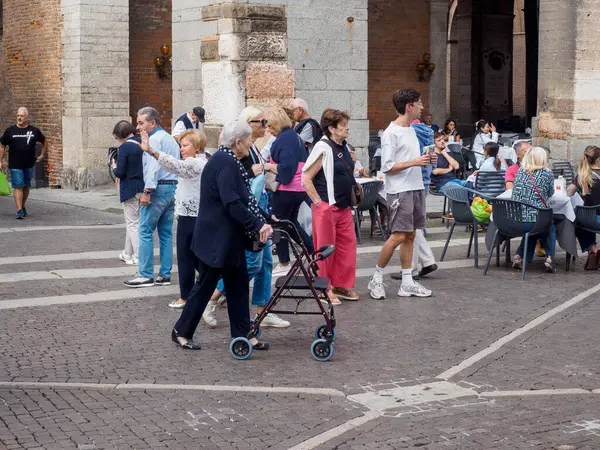 Cremona, Italy - September 18th 2025 Elderly couple with walking frame navigates a cobblestone square near historic Italian architecture with people socializing at outdoor cafe tables enjoying a warm sunny afternoon in Lombardy