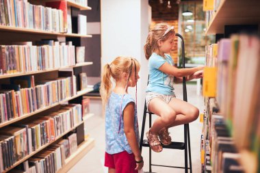 Okul kütüphanesinde okumak için kitap arayan liseli kızlar. Öğrenciler kitap seçiyor. İlköğretim. Ev ödevi. Kitaplardan öğreniyorum. Okula geri dön.