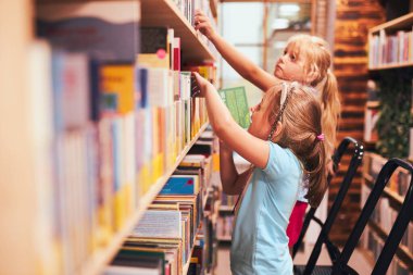 Okul kütüphanesinde kitap arayan liseli kızlar. Belirlenmiş kitapları seçen öğrenciler. İlköğretim. Ev ödevi. Kitaplardan öğreniyorum. Okula geri dön.