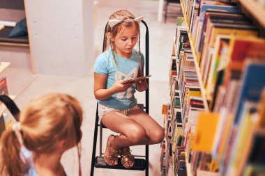 Okul kütüphanesinde sesli kitap arayan liseli kızlar. Belirlenmiş kitapları seçen öğrenciler. İlköğretim. Ev ödevi. Kitaplardan öğreniyorum. Okula geri dön.