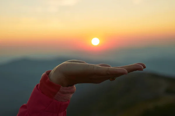 Sun over female hand. Sunrise in mountains. Ready for adventure. Natural mountain landscape with illuminated misty peaks, foggy slopes and valleys, blue sky with orange yellow sunlight