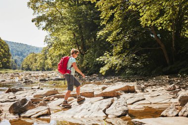 Sırt çantası konsept görüntüsüyle seyahat. Yürüyüş botu giyen sırt çantalı bir kadın dağ nehrinden geçiyor. Yaz gezisi sırasında dağlarda yürüyüş yapan bir kadın. Tatil gezisi doğaya yakın. Doğal manzara.