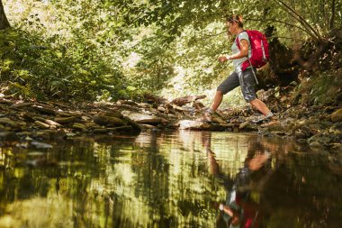 Sırt çantası konsept görüntüsüyle seyahat etmek. Sırt çantalı kadın, dağ nehrinden geçen çizmeli. Yaz gezisi sırasında dağlarda yürüyüş yapan bir kadın. Tatil gezisi doğaya yakın. Doğal manzara.