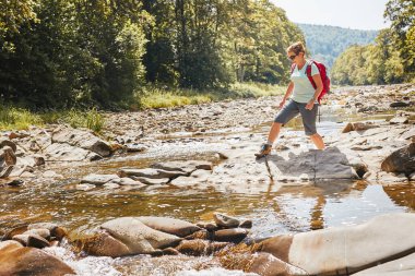 Sırt çantası konsept görüntüsüyle seyahat. Yürüyüş botu giyen sırt çantalı bir kadın dağ nehrinden geçiyor. Yaz gezisi sırasında dağlarda yürüyüş yapan bir kadın. Tatil gezisi doğaya yakın. Doğal manzara.