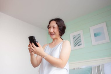 Evde cep telefonu kullanan mutlu Asyalı kadın. Asyalı ev hanımı akıllı telefondan sosyal medyayı kontrol ediyor. Sakin ol kadın. Cep telefonu uygulaması kullanıyor. Sipariş teslimatı yapıyor, internetten alışveriş yapıyor.