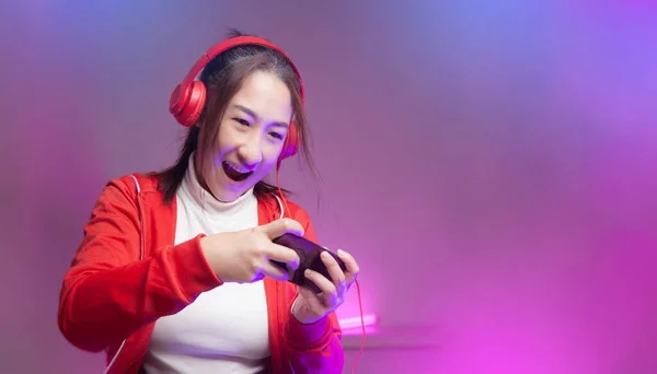 Kulaklıklı mutlu Asyalı bayan oyuncu akıllı telefondan oyun oynuyor. Profesyonel flamalar kırmızı giysiler içinde oyun oynuyorlar sosyal oyun oynarken eğleniyorlar. Siber Neon Işıkları Biçimi