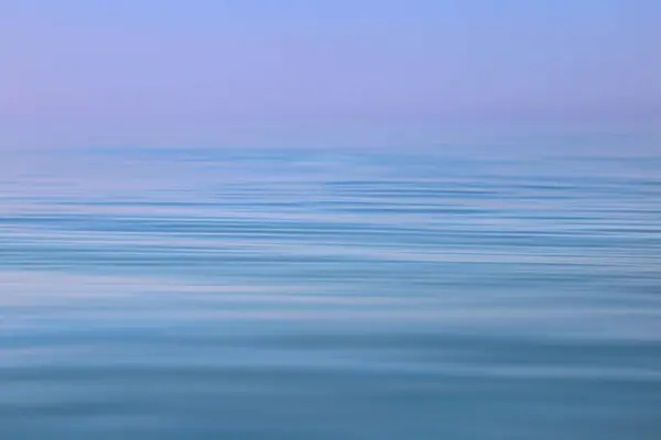 Açık bir gökyüzünün altında denizin dalgalı sakin yüzeyi. Mavi ve mor arkaplan.
