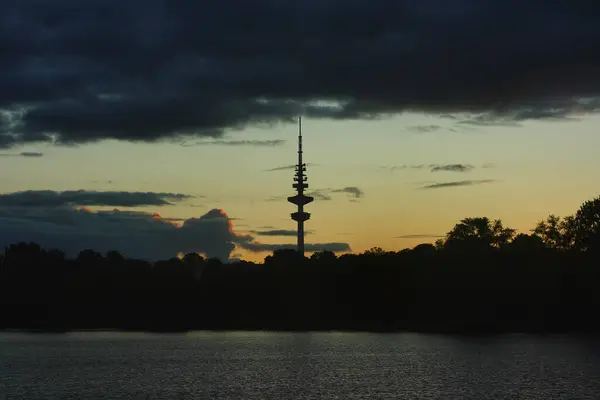 Hamburg TV kulesi. Gölün su yüzeyi ve Hamburg TV kulesinin çevresi bulutlardan oluşan bir şerit ve şehir parkının kara hatlarıyla akşam gökyüzüne karşı..