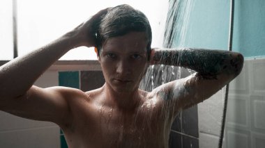 Genç adam saçlarını ve vücudunu yıkıyor, banyoda suyun altında duruyor. Yüksek kalite fotoğraf
