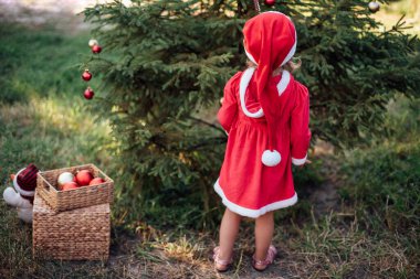 Temmuz 'da Noel. Yazın Noel 'i ormanda bekleyen bir çocuk. Noel ağacı süsleyen kırmızı elbiseli küçük kızın portresi. Kış tatili ve insanların konsepti. Mutlu Noeller ve mutlu tatiller