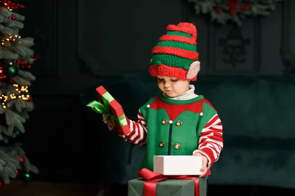 Merry Christmas and Happy Holidays. Surprised cute child boy opening present. Little kid having fun near Christmas tree indoors. Funny kid in Santa hat with gift box near christmas tree