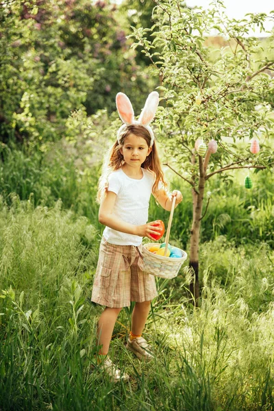 Paskalya yumurtası avı. Bahçede yumurta toplamak için koşan tavşan kulaklı bir kız çocuğu. Paskalya geleneği. Sepeti renkli yumurtalarla dolu bir bebek. geniş açı görünümü