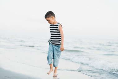 Denizde yaz tatili. Deniz kenarında çizgili tişörtlü bir çocuk. Çocuk yaz tatilinde sahilde koşuyor ve eğleniyor. Mutlu çocuk gün batımında oynuyor. Mutlu bir çocukluk. Seyahat ve macera konsepti