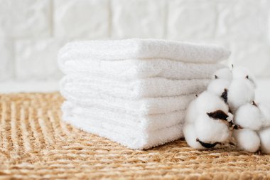 Beyaz pamuklu örülmüş hasırın ve pamuğun üzerinde beyaz havlu yığını. Beyaz desenli duvar. Lüks, rahatlama, kişisel bakım, ev dekorasyonu, spa ve sağlık kavramı..