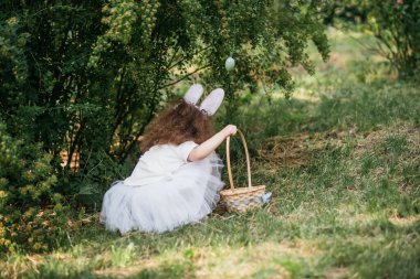 Paskalya yumurtası avı. Bahçede yumurta toplamak için koşan tavşan kulaklı bir kız çocuğu. Paskalya geleneği. Renkli yumurtalarla dolu sepeti olan bebek.