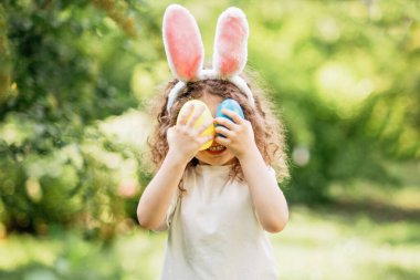 Paskalya yumurtası avı. Bahçede yumurta toplamak için koşan tavşan kulaklı bir kız çocuğu. Paskalya geleneği. Renkli yumurtalarla dolu sepeti olan bebek.
