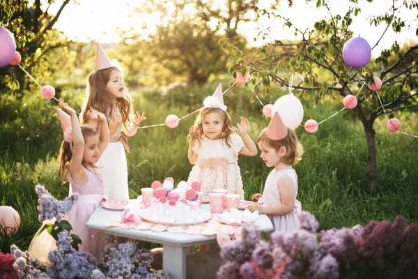 夏季户外儿童生日派对 一群快乐的孩子在公园里庆祝生日 孩子们在生日蛋糕上放蜡烛 孩子们举行紫丁香粉红乳胶装饰和食物派对 礼物和糖果 图库图片