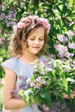 Kıvırcık sarı saçlı genç kız güneşli bahçede çiçek açan leylak çalılarının arasında duruyor. yumuşak ışıklandırma ve doğal ortam rüya gibi bir atmosfer yaratır, masumiyet ve çocukluk temaları uyandırır.,