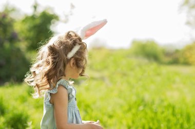 Paskalya yumurtası avı. Bahçede yumurta toplamak için koşan tavşan kulaklı bir kız çocuğu. Paskalya geleneği. Renkli yumurtalarla dolu sepeti olan bebek.