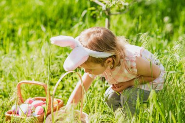 Paskalya yumurtası avı. Bahçede yumurta toplamak için koşan tavşan kulaklı bir kız çocuğu. Paskalya geleneği. Renkli yumurtalarla dolu sepeti olan bebek.