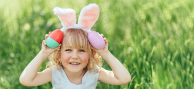 Paskalya yumurtası avı. Bahçede yumurta toplamak için koşan tavşan kulaklı bir kız çocuğu. Paskalya geleneği. Renkli yumurtalarla dolu sepeti olan bebek.