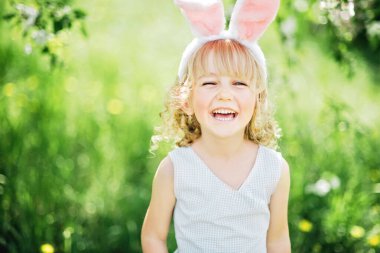 Paskalya yumurtası avı. Bahçede yumurta toplamak için koşan tavşan kulaklı bir kız çocuğu. Paskalya geleneği. Renkli yumurtalarla dolu sepeti olan bebek.