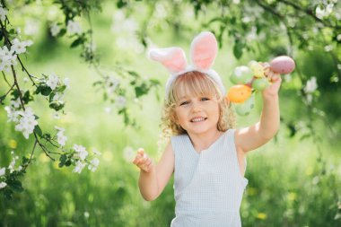 Paskalya yumurtası avı. Bahçede yumurta toplamak için koşan tavşan kulaklı bir kız çocuğu. Paskalya geleneği. Renkli yumurtalarla dolu sepeti olan bebek.