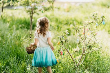 Paskalya yumurtası avı. Bahçede yumurta toplamak için koşan tavşan kulaklı bir kız çocuğu. Paskalya geleneği. Renkli yumurtalarla dolu sepeti olan bebek.