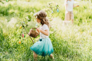 Paskalya yumurtası avı. Bahçede yumurta toplamak için koşan tavşan kulaklı bir kız çocuğu. Paskalya geleneği. Renkli yumurtalarla dolu sepeti olan bebek.