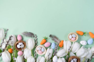 Creative Easter flat lay featuring white tulips, speckled pastel eggs in nests, artificial carrots, pussy willow branches, and soft feathers on a mint green background.