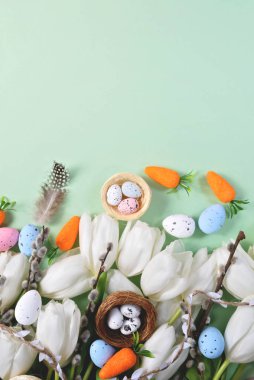 Creative Easter flat lay featuring white tulips, speckled pastel eggs in nests, artificial carrots, pussy willow branches, and soft feathers on mint green background. space for festive advertisements