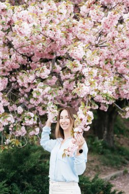 Gülümseyen kadın çiçek açan pembe kiraz ağaçlarının arasında açık mavi bir gömlek giyerek ve açık havada güneşli bir bahar gününün tadını çıkararak duruyor.