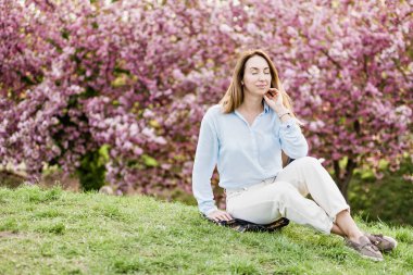 Gülümseyen kadın çiçek açan pembe kiraz ağaçlarının arasında açık mavi bir gömlek giyerek ve açık havada güneşli bir bahar gününün tadını çıkararak duruyor.