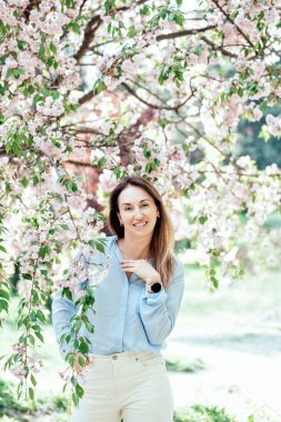 Gülümseyen kadın çiçek açan pembe kiraz ağaçlarının arasında açık mavi bir gömlek giyerek ve açık havada güneşli bir bahar gününün tadını çıkararak duruyor.