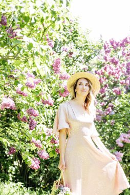 Yazlık Şapkalı Genç Kadın güneşli bir günde leylak çiçeklerinin kokusunun tadını çıkarıyor. Hasır şapkalı ve elbiseli bayan bahar güneşli bahçede leylak çiçekleri kokluyor..