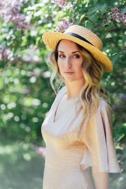 Yazlık Şapkalı Genç Kadın güneşli bir günde leylak çiçeklerinin kokusunun tadını çıkarıyor. Hasır şapkalı ve elbiseli bayan bahar güneşli bahçede leylak çiçekleri kokluyor..