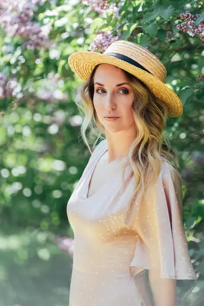 Yazlık Şapkalı Genç Kadın güneşli bir günde leylak çiçeklerinin kokusunun tadını çıkarıyor. Hasır şapkalı ve elbiseli bayan bahar güneşli bahçede leylak çiçekleri kokluyor..