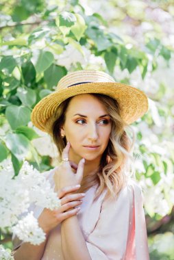 Yazlık Şapkalı Genç Kadın güneşli bir günde leylak çiçeklerinin kokusunun tadını çıkarıyor. Hasır şapkalı ve elbiseli bayan bahar güneşli bahçede leylak çiçekleri kokluyor..