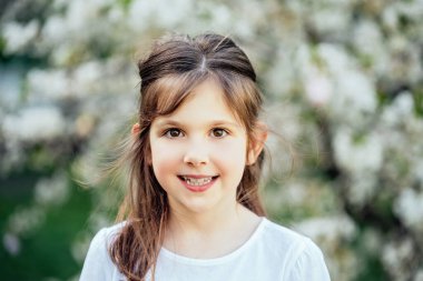 Çiçek açan ağaçlar arasındaki neşeli çocukluk anı. Kahverengi saçlı, beyaz tişörtlü gülümseyen genç kız çiçek açan bahar ağaçlarının önünde dikiliyor.