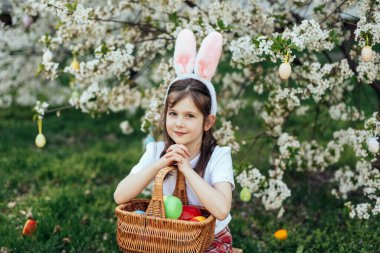 Paskalya yumurtası avı. Bahçede yumurta toplamak için koşan tavşan kulaklı bir kız çocuğu. Paskalya geleneği. Renkli yumurtalarla dolu sepeti olan bebek.