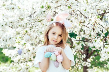 Paskalya yumurtası avı. Bahçede yumurta toplamak için koşan tavşan kulaklı bir kız çocuğu. Paskalya geleneği. Renkli yumurtalarla dolu sepeti olan bebek.