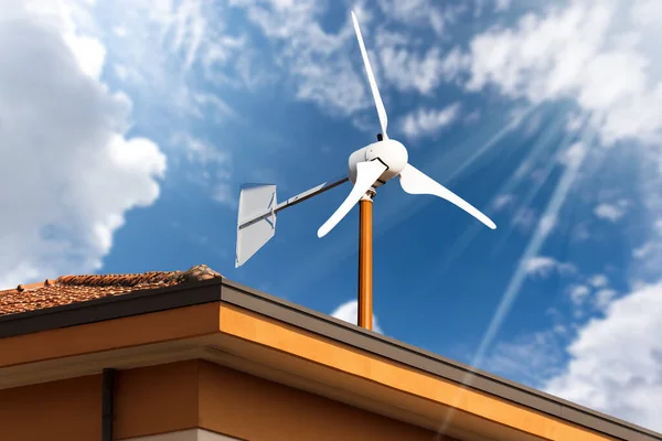 stock image Close-up of a small wind turbine on the top of a roof of a house, against a blue sky with clouds and sunbeams. Renewable energy concept.