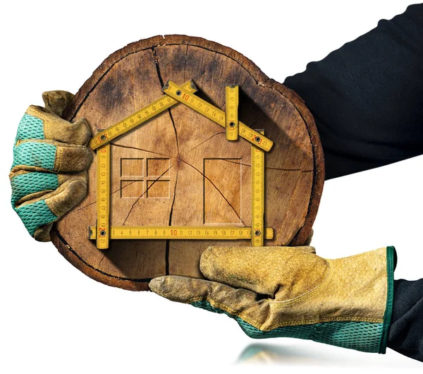 白い背景に隔離された家の形で黄色の木製の折りたたみ定規と木の幹の断面を保持保護作業手袋を持つ大工 — ストック写真