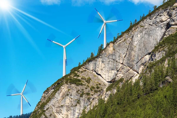 山の岩の顔の上に白と青の風力タービンのグループは 背景に澄んだ青い空とサンビーム 手前の松林 再生可能エネルギーの概念 — ストック写真
