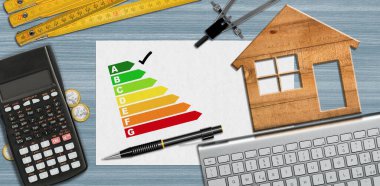 Ev enerjisi verimliliği derecesi. Masasında enerji verimliliği grafiği, hesap makinesi, katlama cetveli, pusula, kalem ve bilgisayar klavyesi olan küçük ahşap bir ev ve kopyalama alanı var..