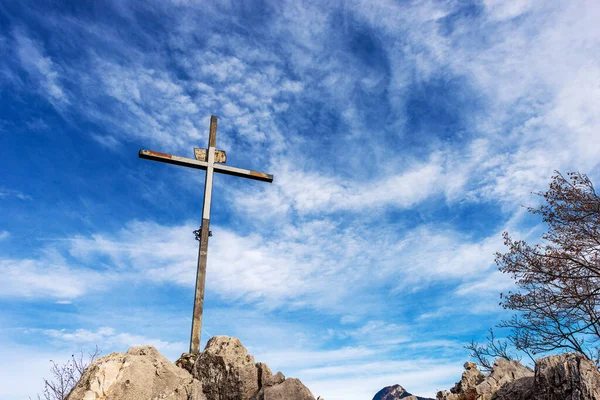 Metal religious cross on the top of a mountain peak against a blue sky with clouds and copy space. Croce di Dos dela Cronela, Monte Altissimo di Nago, Trentino Alto Adige, Italy, Europe.