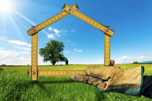 Design or project concept of an ecological house. Hand with work glove holding a folding ruler in the shape of a house, on a green meadow with a green tree. Clear blue sky and sunbeams on background.